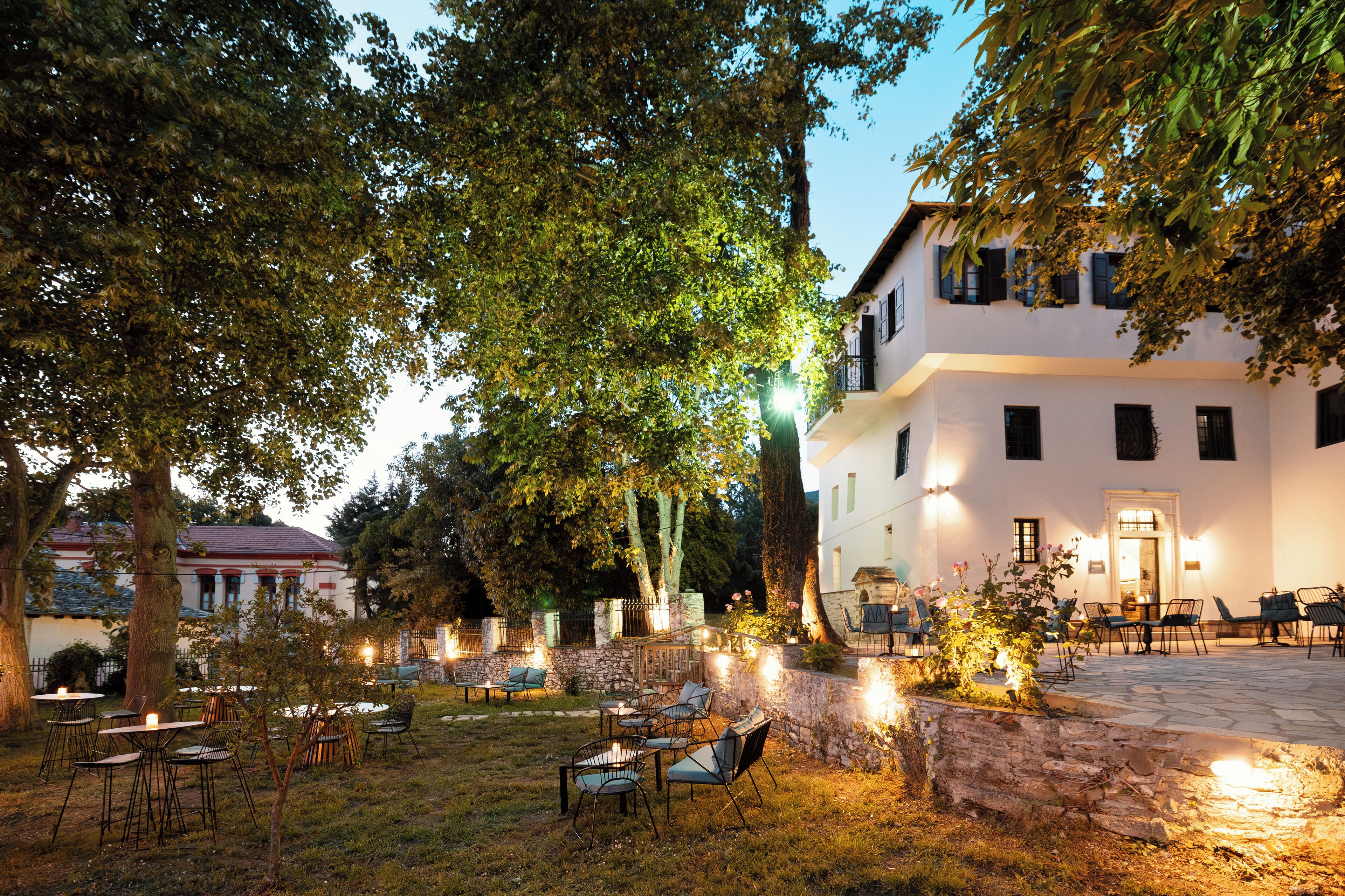 An outdoor seating area with tables and chairs, surrounded by trees and a stone wall.