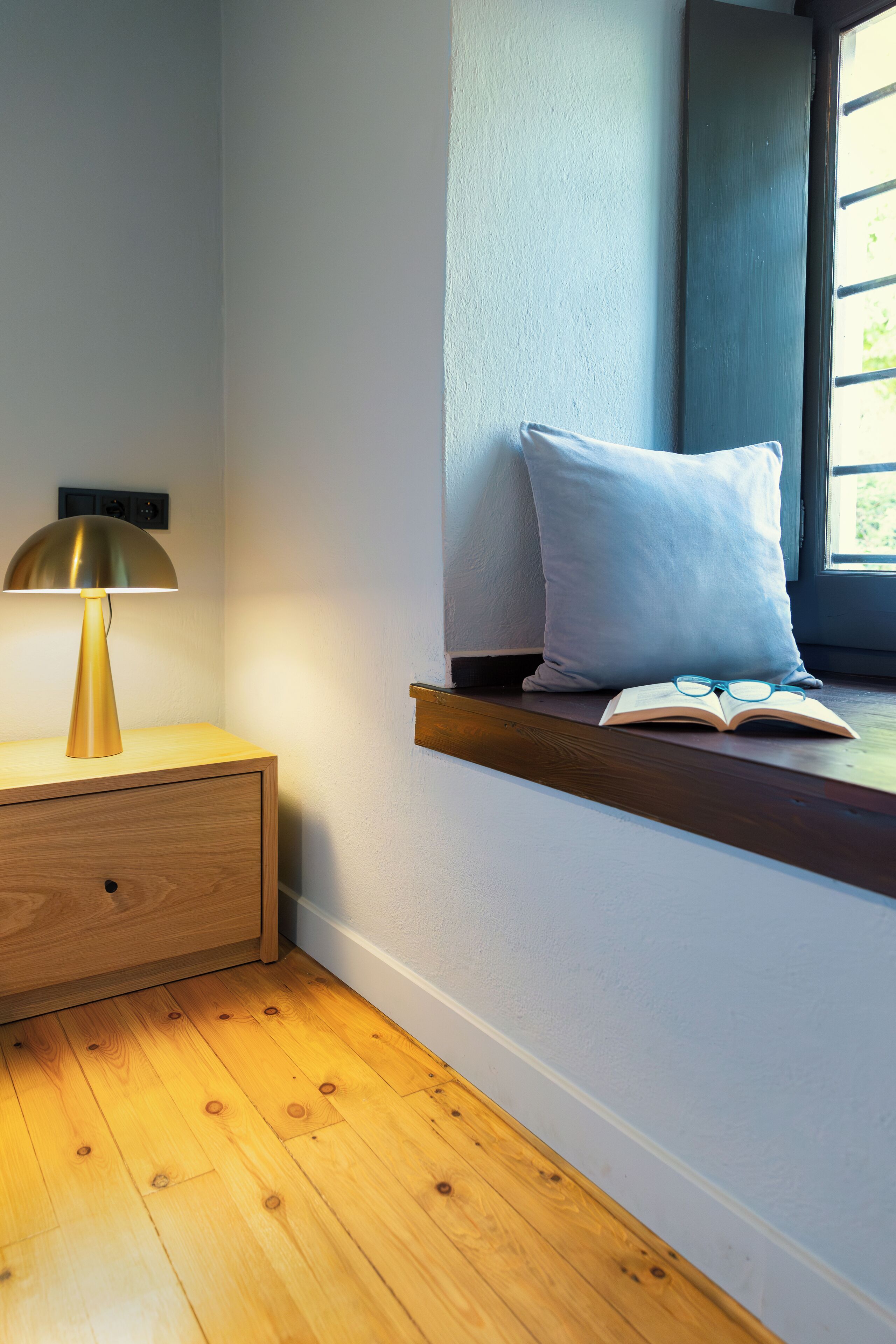 A wooden bedside table with a lamp, a pillow, and an open book on a window sill.