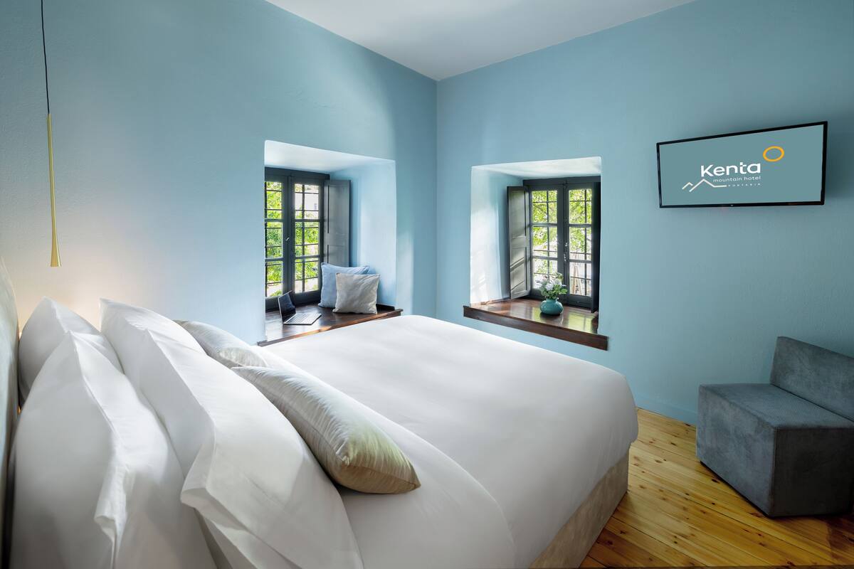 A modern hotel room with a large bed, a small seating area, and a TV displaying the hotel name.