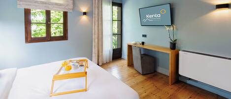 A hotel room with a bed, a desk, a chair, a window with curtains, a wall-mounted TV, and a radiator.