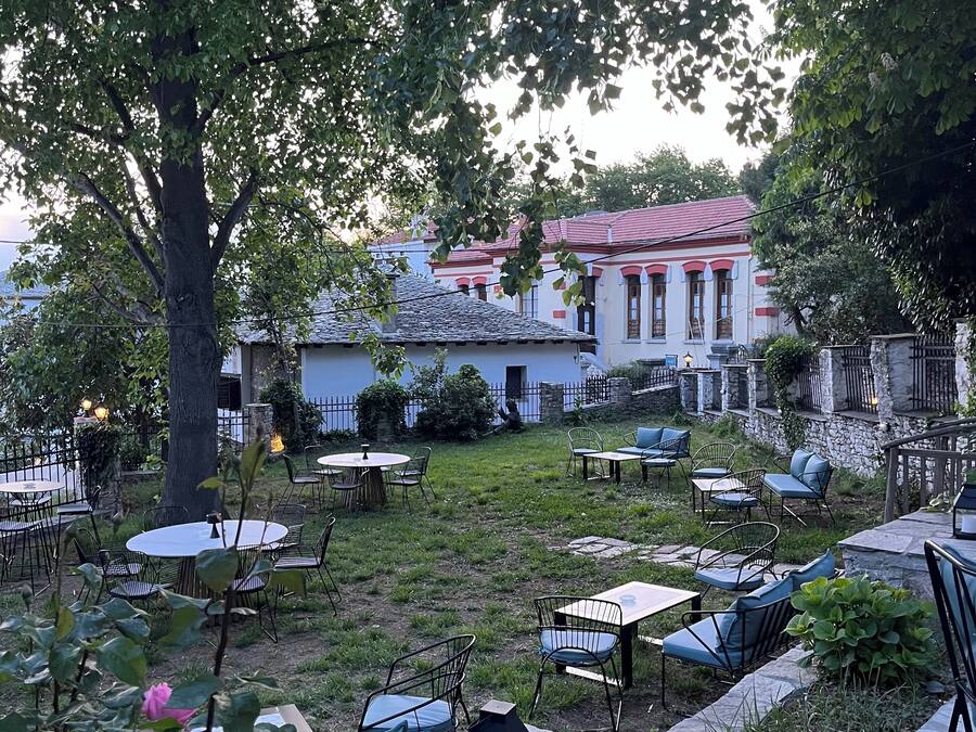 An outdoor seating area with tables and chairs, surrounded by greenery and a stone wall.