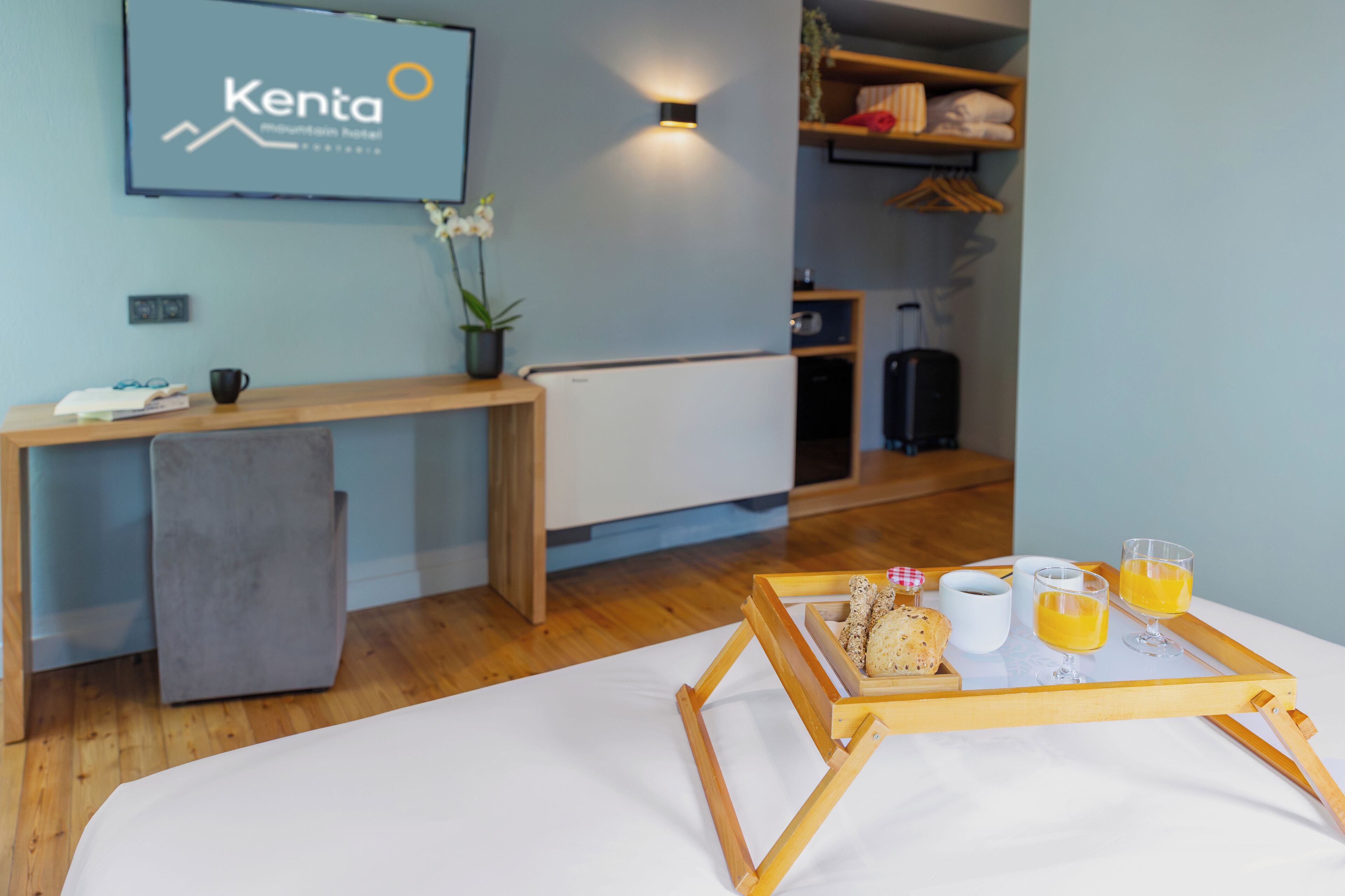 Hotelkamer met een bed, bureau, kleine koelkast, een dienblad met ontbijtartikelen en een wandgemonteerde televisie waarop de naam van het hotel staat.