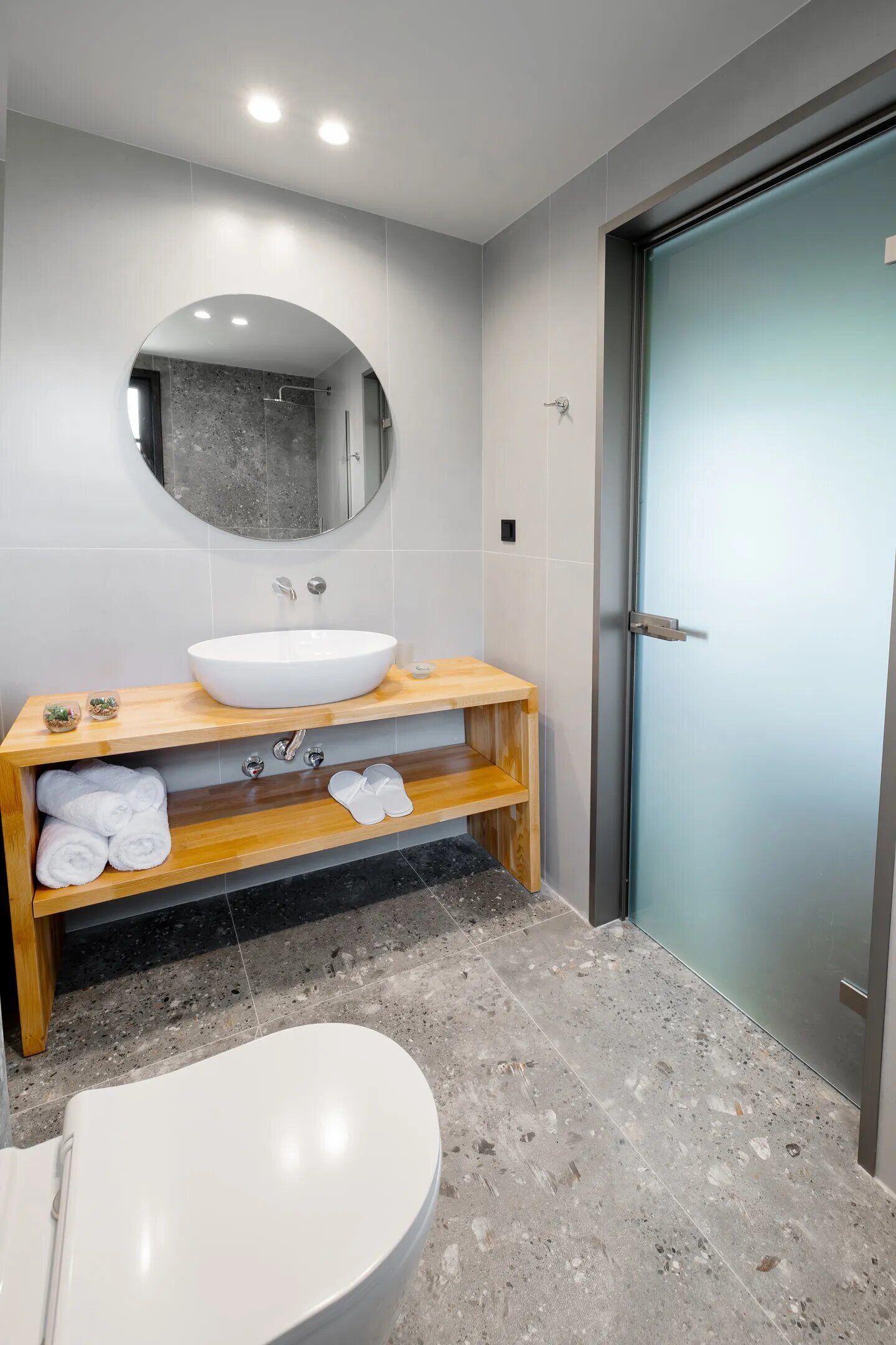 Une salle de bain moderne avec un lavabo blanc, un meuble-lavabo en bois, un miroir rond et une porte en verre.
