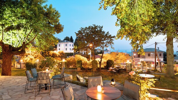 An outdoor patio with stone flooring, round tables, and chairs, surrounded by trees and a view of buildings in the background. - Kenta Mountain Hotel (Volos)