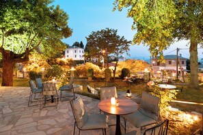 An outdoor patio with stone flooring, round tables, and chairs, surrounded by trees and a view of buildings in the background.