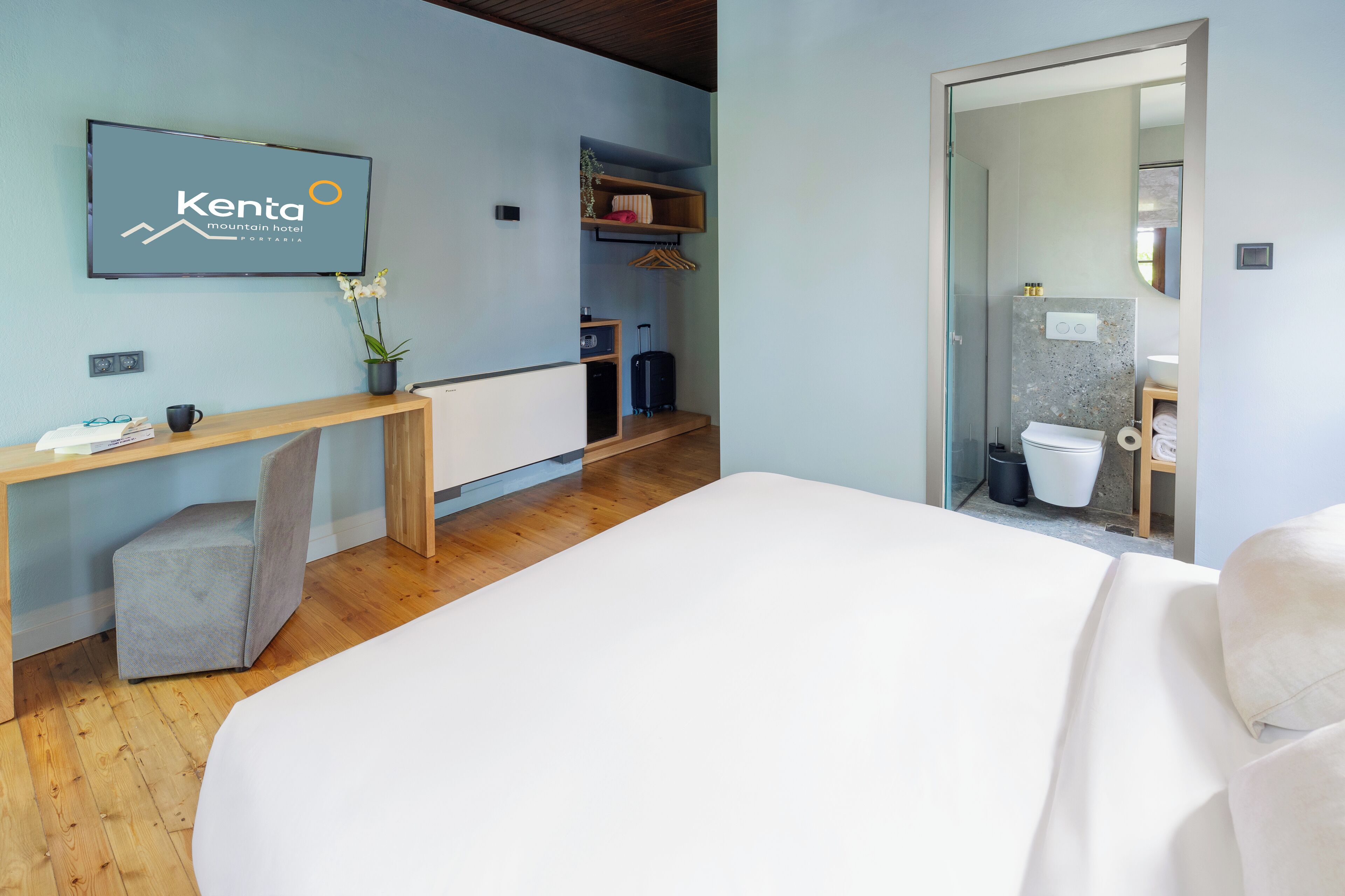 A hotel room with a bed, a desk with a computer, a TV mounted on the wall, a small plant, and a suitcase.