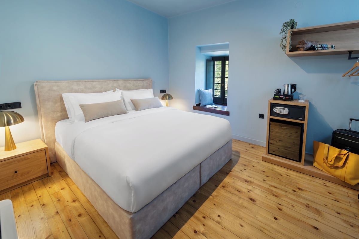 A modern bedroom with a large bed, bedside tables, a wooden floor, a small desk with a radio, and a window with a view.