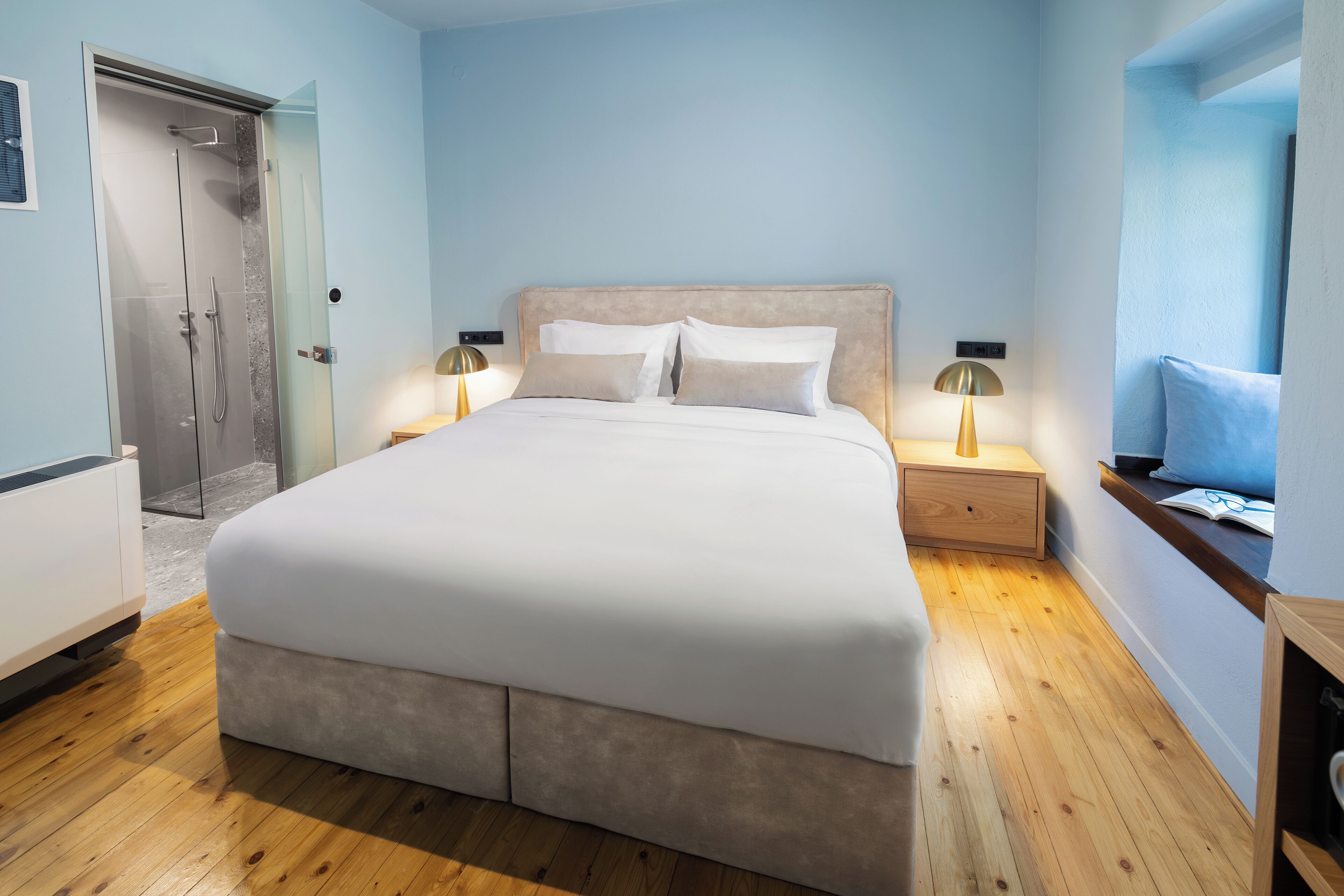 A modern bedroom with a large bed, bedside lamps, a wooden floor, and a glass shower enclosure.