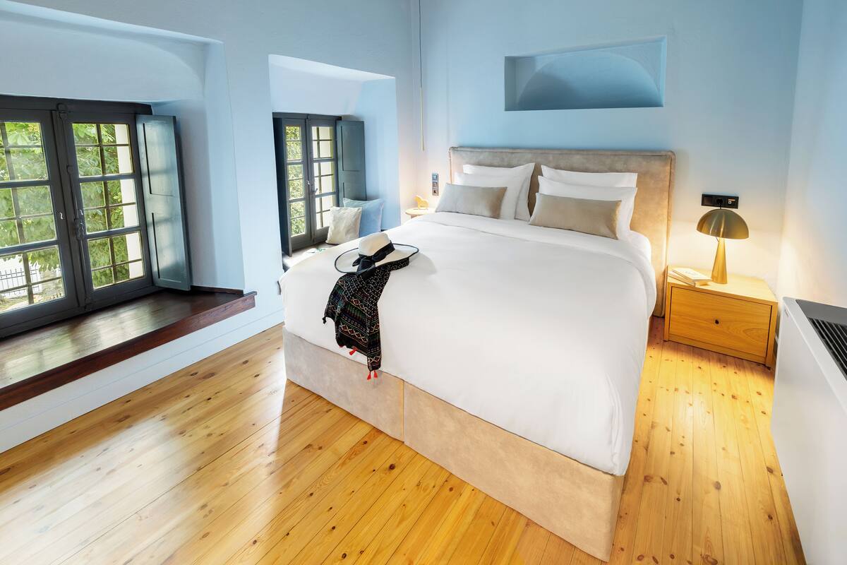 A modern bedroom with a large bed, wooden flooring, and a view of greenery through the windows.
