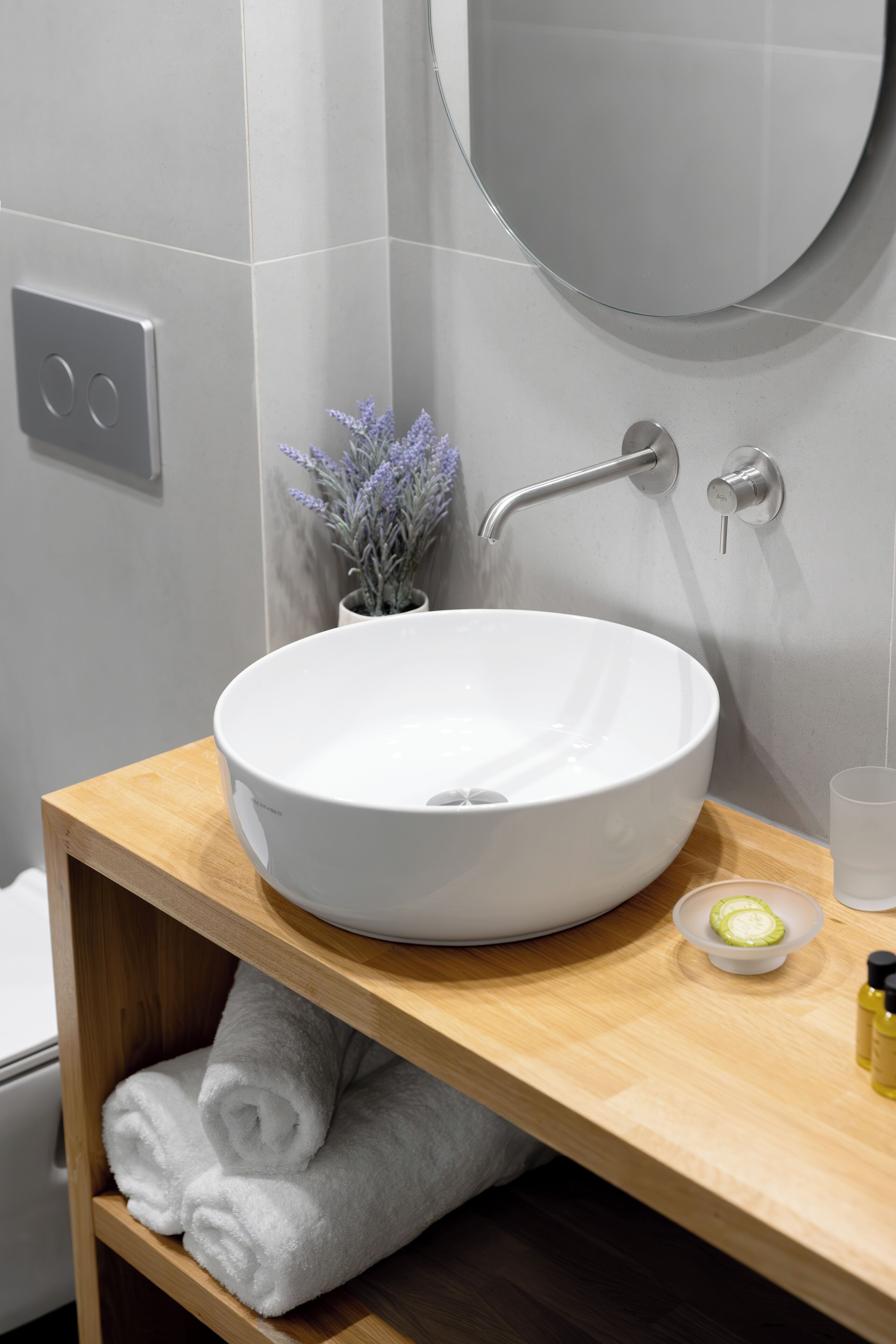 A modern bathroom with a white vessel sink, a wooden vanity, a small potted lavender plant, and a glass of water.