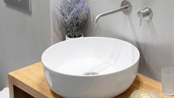 A modern bathroom with a white vessel sink, a wooden vanity, a small potted lavender plant, and a glass of water.