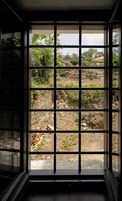 A window with a grid pattern, through which a view of a rural landscape with trees and a stone wall is visible.