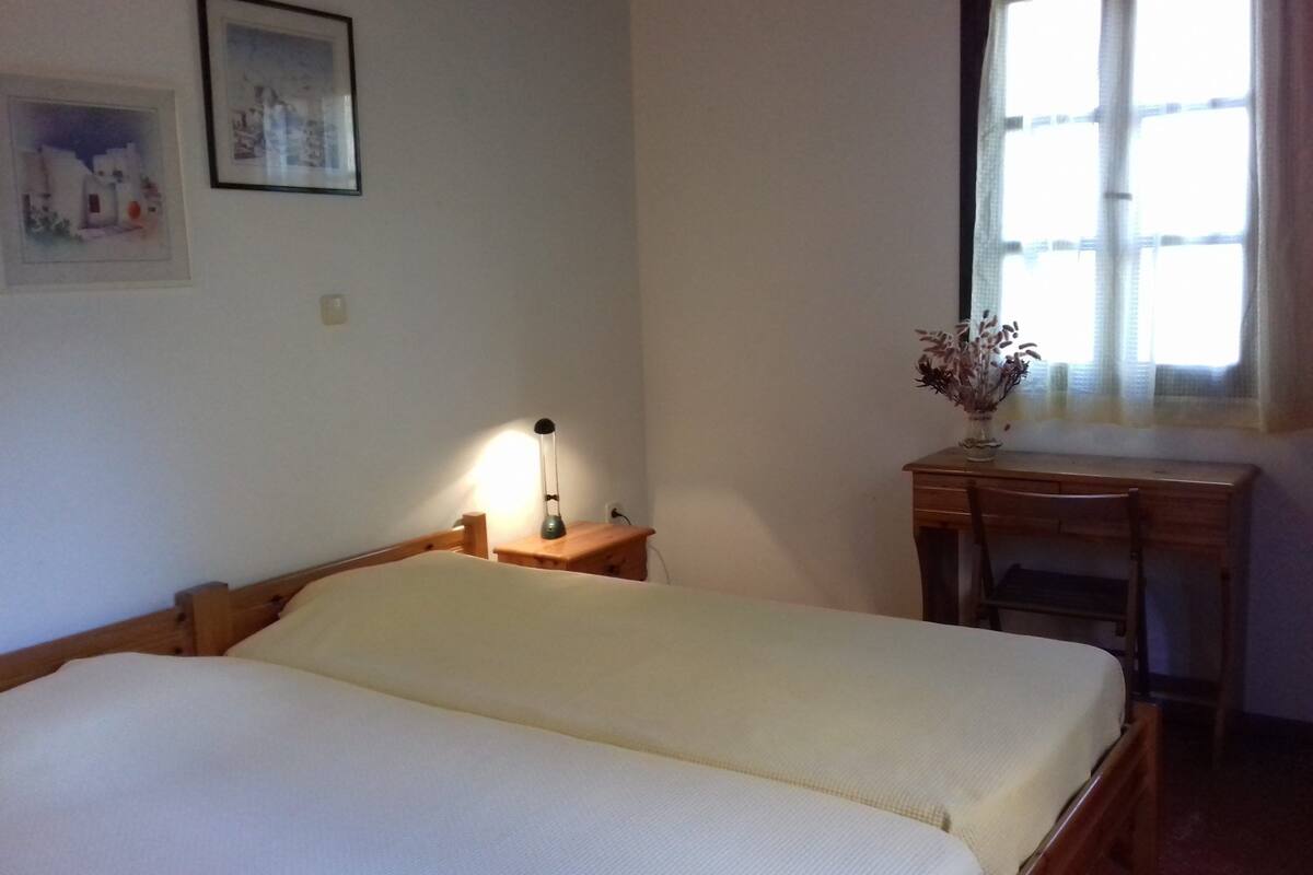 A bedroom with two single beds, a bedside table with a lamp, a desk with a chair, and a window with curtains.