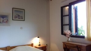Un dormitorio con cama, escritorio, silla, lámpara y ventana con vista a follaje.