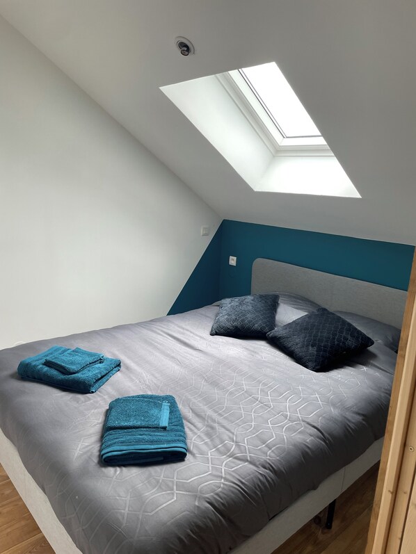 Ein Schlafzimmer mit Dachfenster, einem Bett mit grauer Bettwäsche, zwei blauen Handtüchern und einer türkisfarbenen Wand.