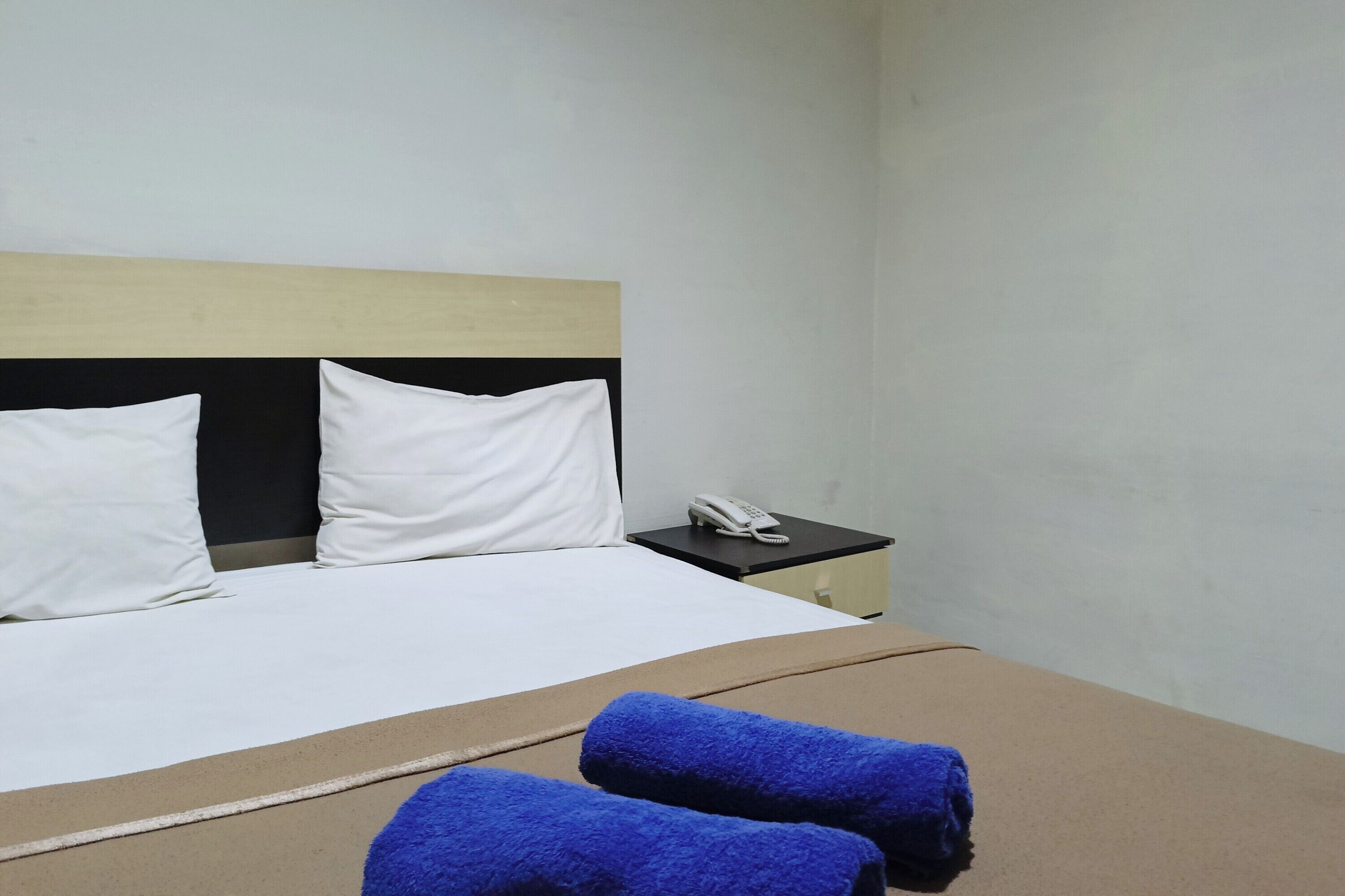 Una camera d'albergo con un letto, due cuscini bianchi, una testiera in legno, un comodino con un telefono bianco e due asciugamani blu.