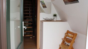 Un couloir avec un escalier en bois menant à une porte, une petite étagère avec une plante et des objets décoratifs, et une chaise en bois.