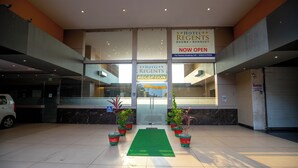 Zona de recepción de hotel con alfombra verde, plantas en macetas y letreros que indican “Hotel Regents Rooms + Banquet” y “Recepción”.