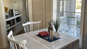 Un coin repas avec une table et des chaises blanches, une plante en pot et une fenêtre donnant sur des structures en pierre à l’extérieur.