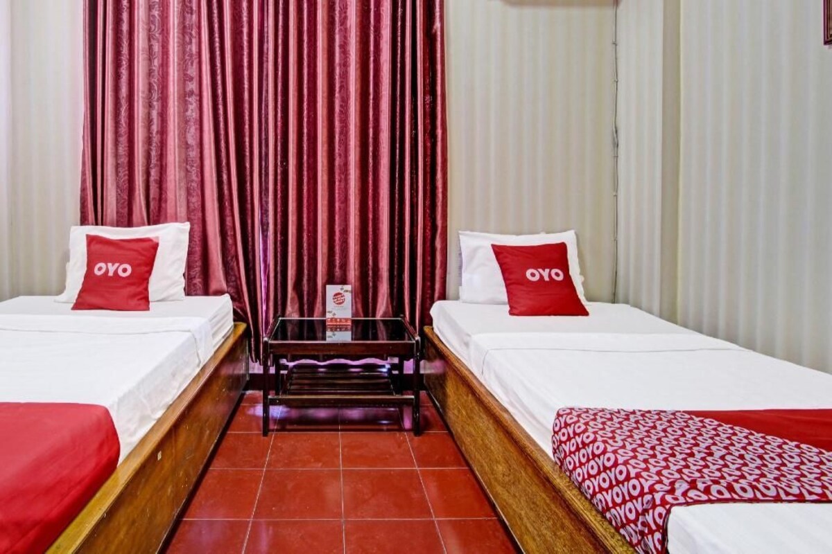 Een hotelkamer met twee bedden, elk met een rood kussen met het 'OYO'-logo, een klein tafeltje in het midden en een rood-wit gebloemd dekbed op een van de bedden.