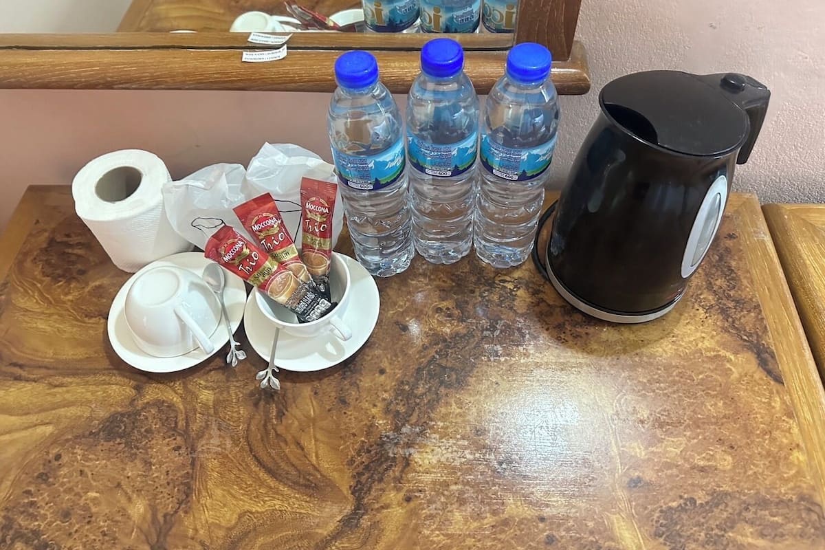 A wooden table with a coffee maker, four bottles of water, a roll of toilet paper, and two cups with coffee packets.