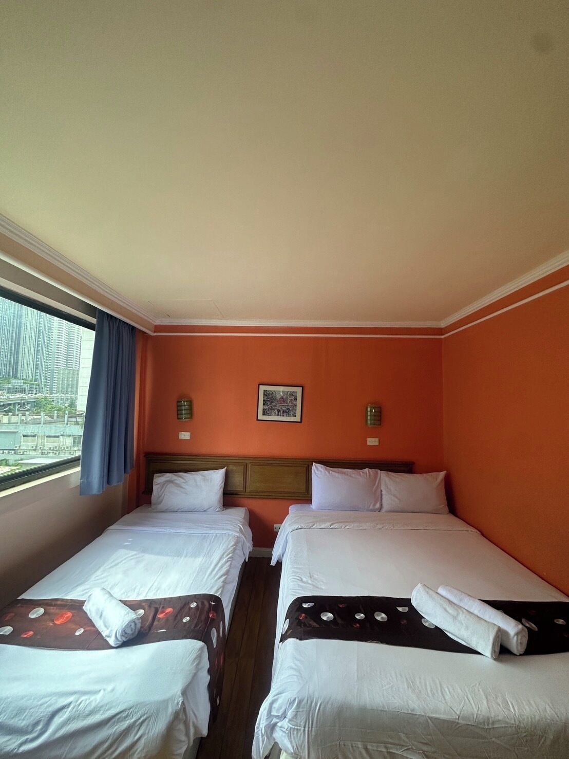 A hotel room with two beds, a window with a view of buildings, and a painting on the wall.
