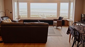 A living room with a large bay window overlooking a beach, a sofa, a table with a patterned tablecloth, and a television. - Apartment in Nieuwpoort With Ocean Views (Nieuwpoort)