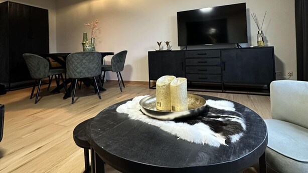 A modern living room with a round black coffee table, a flat-screen TV mounted on the wall, a dining area with a dark wooden cabinet and grey chairs, and a light-colored sofa. - Maila - Marbi (Varel)