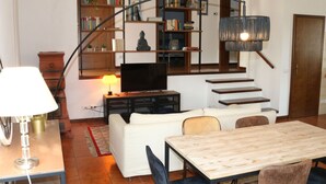 Ein modernes Wohnzimmer mit einer Couch, einem Esstisch und einem Fernseher. Eine Treppe mit Bücherregalen und einer Statue ist ebenfalls zu sehen.