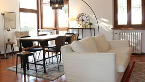 Ein modernes Wohnzimmer mit einem weißen Sofa, einem Esstisch mit Stühlen, einer großen Uhr und einem Kronleuchter.