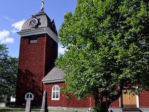 Uma igreja de tijolos vermelhos com uma torre de relógio, uma grande árvore na frente e um céu azul claro.