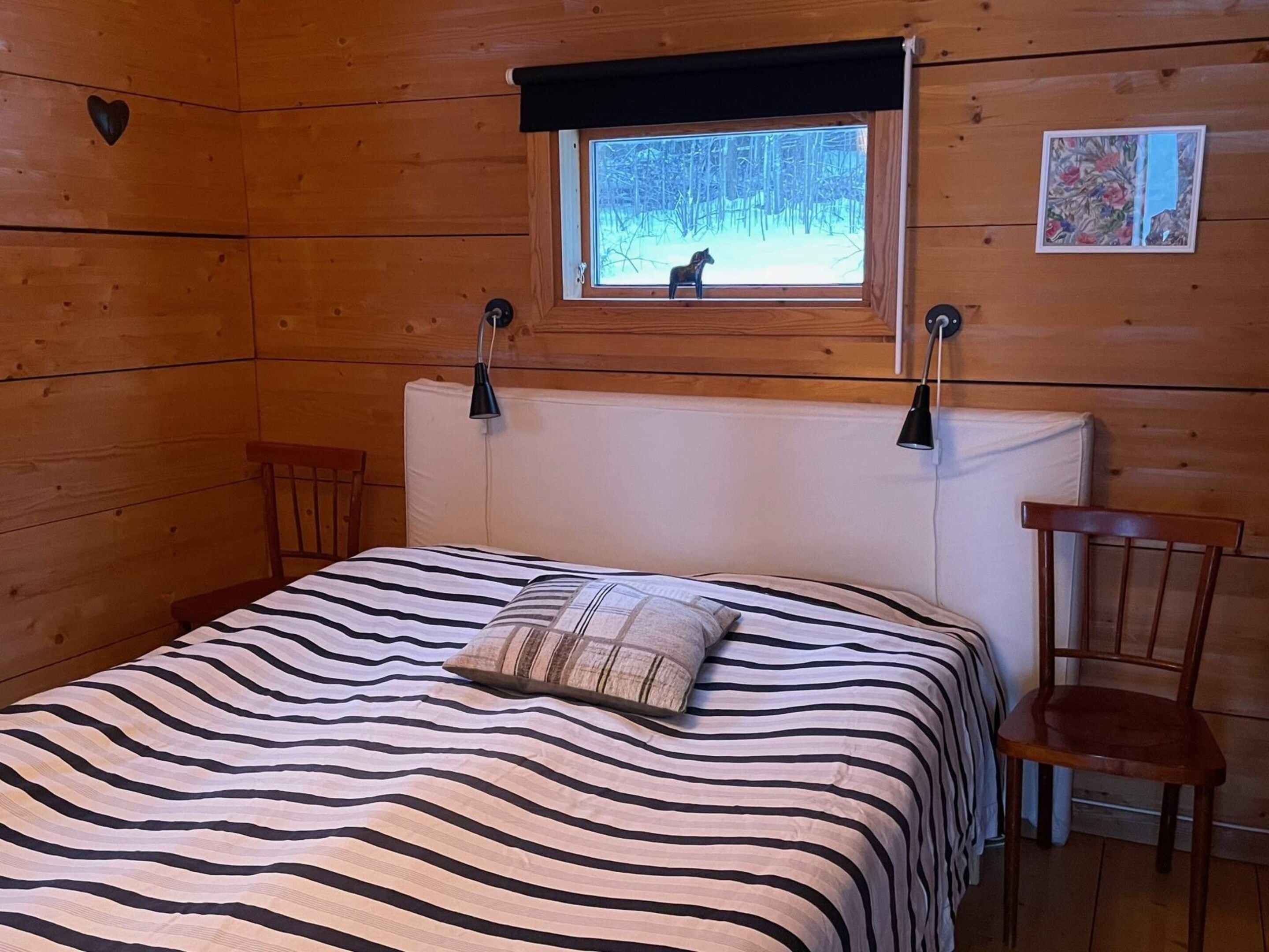 A wooden room with a bed, a window with a view of trees, a chair, and a wall-mounted lamp.