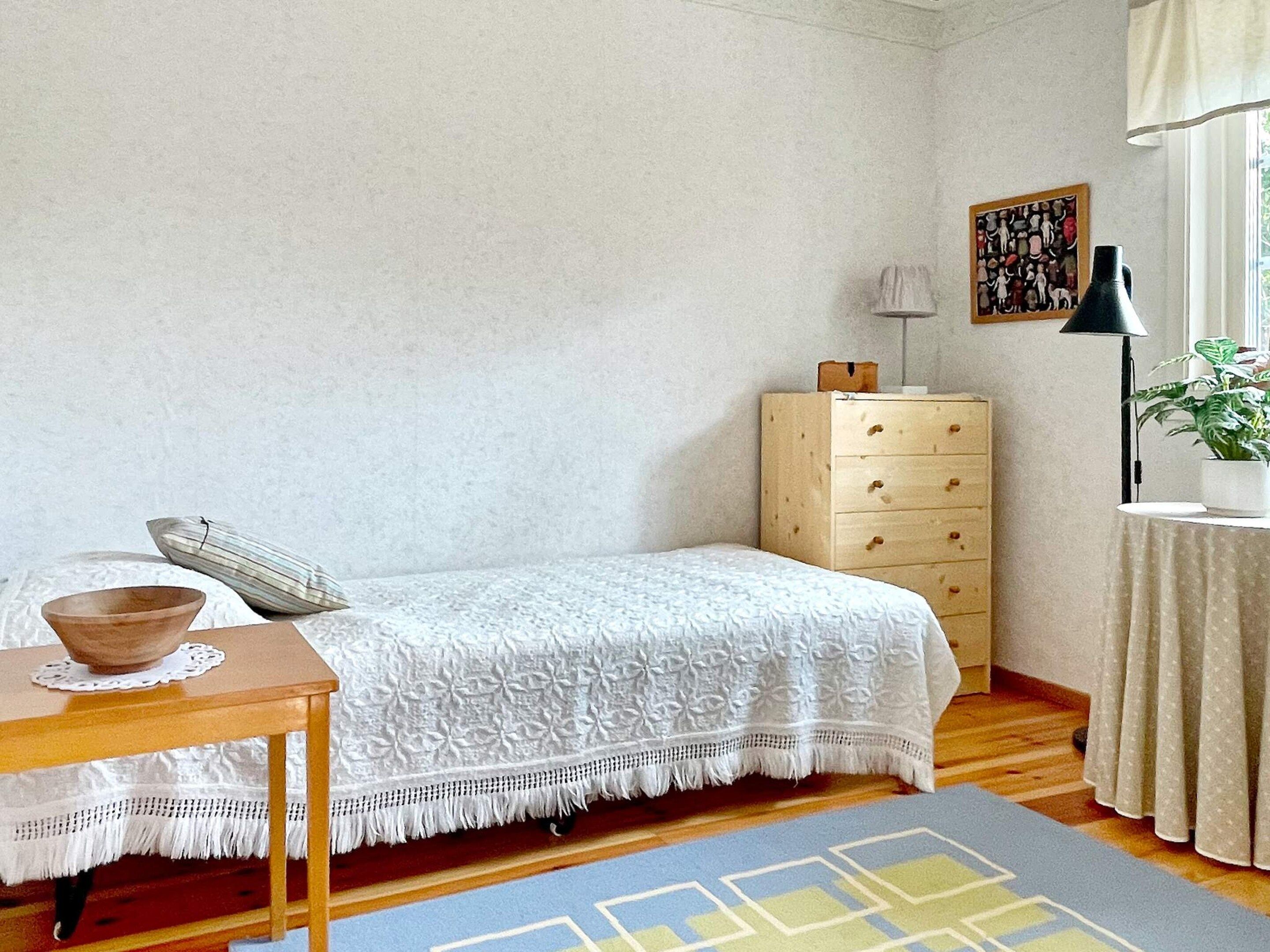 Quarto com cama, cômoda de madeira, mesa pequena com tigela, luminária e planta.