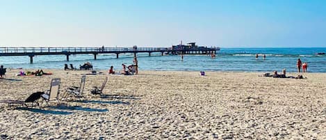 A beach with a pier extending into the water, sun loungers, and people enjoying the seaside.