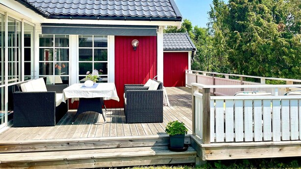 Una terraza de madera con mobiliario exterior, un edificio rojo con techo negro y una valla blanca.