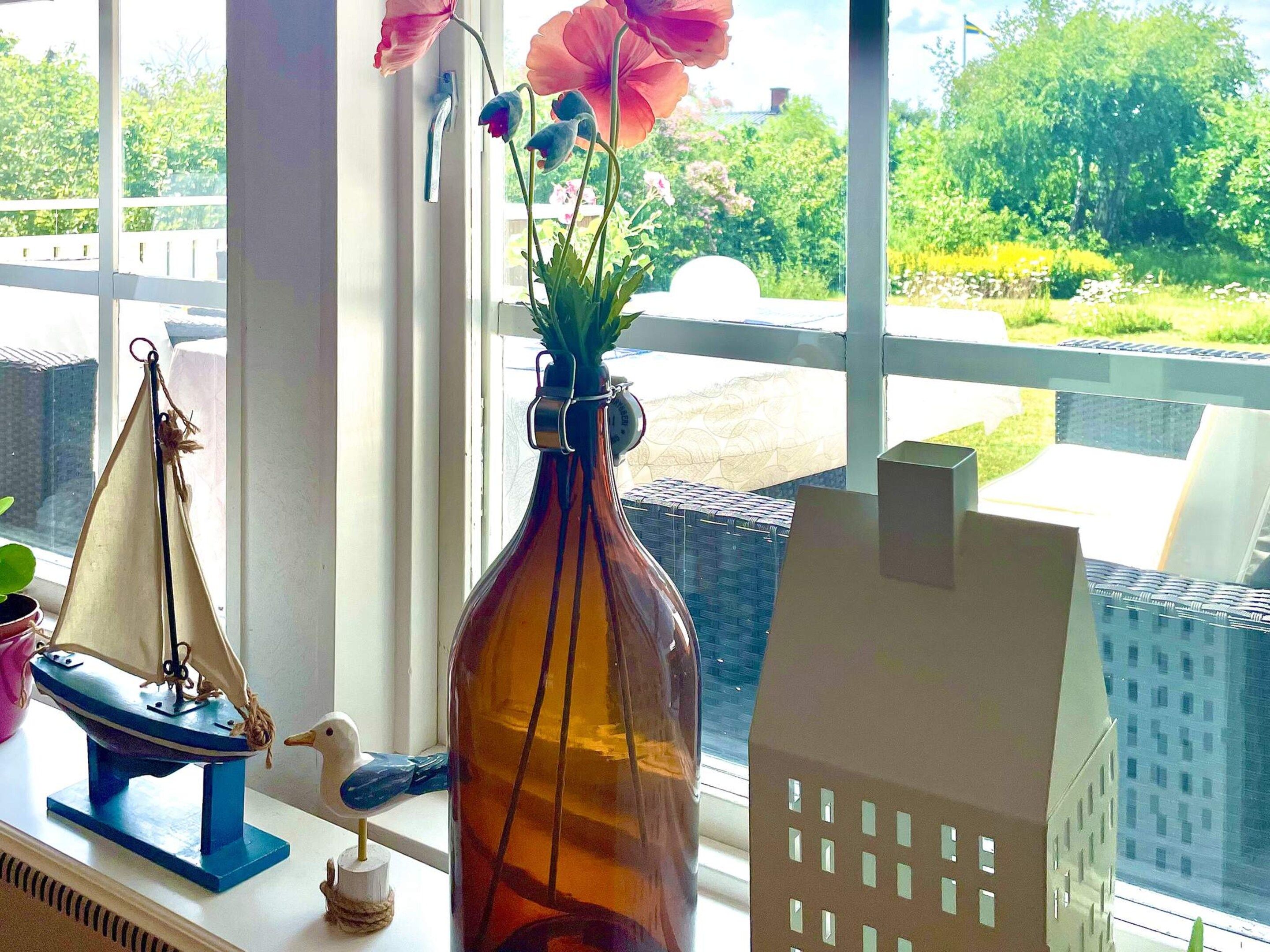 A window with a view of greenery and a building, a vase with flowers, a model sailboat, and a small bird figurine.