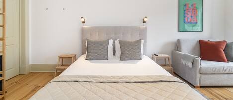 A bedroom with a large bed, two bedside tables, a grey sofa, and a colorful abstract painting on the wall.