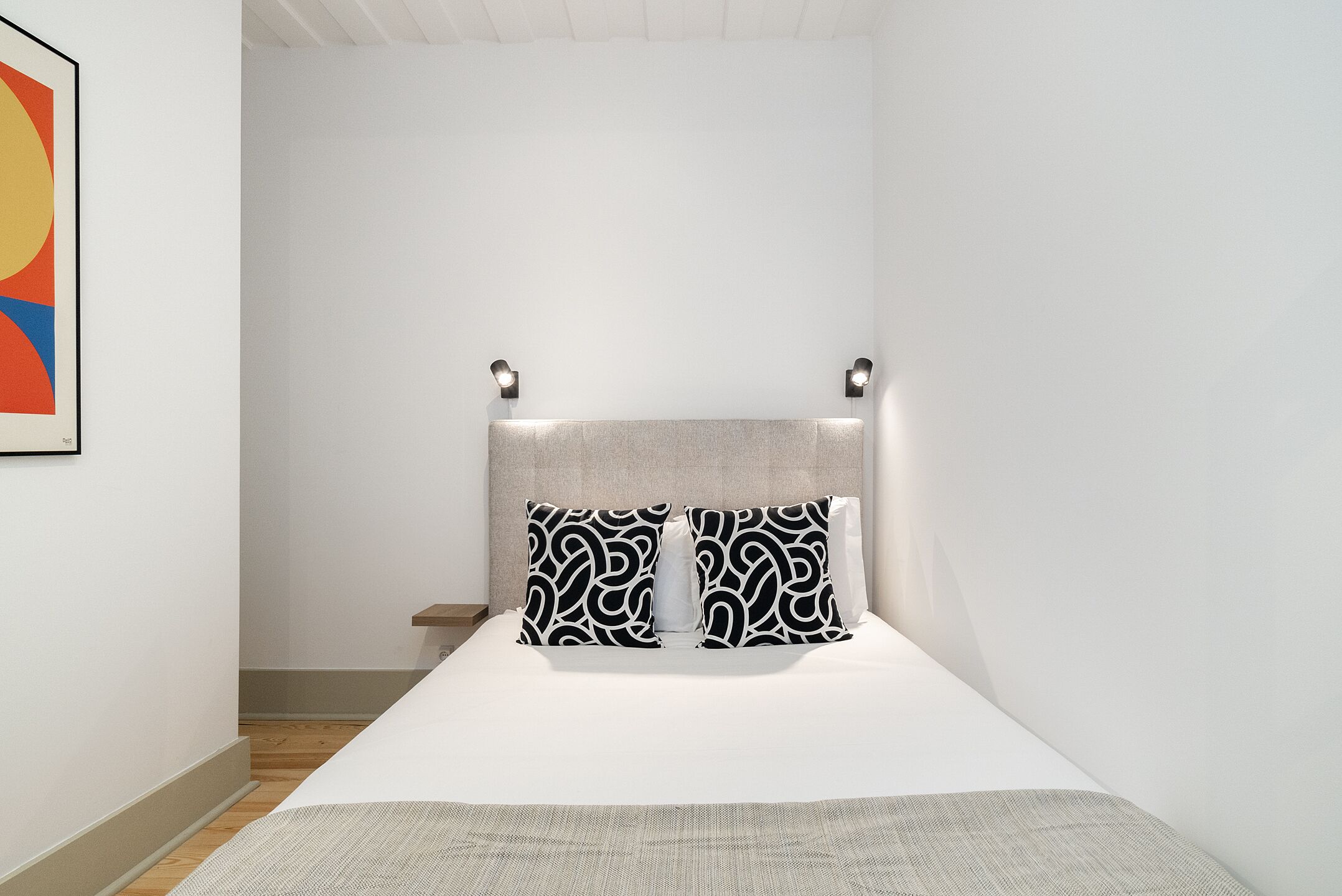 A minimalist bedroom with a bed, two wall-mounted lights, and a single artwork on the left wall.