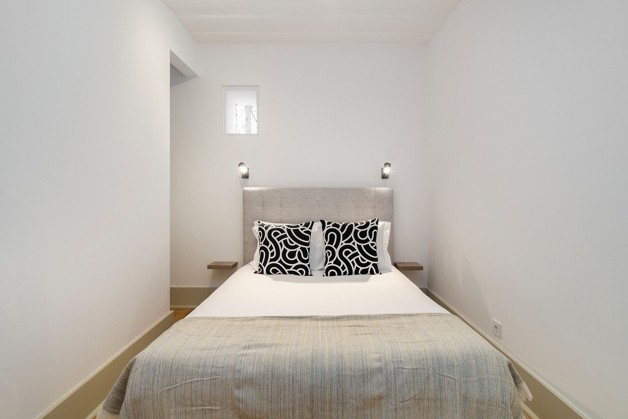 Une chambre d’hôtel étroite, avec un lit, deux oreillers, une tête de lit et une petite fenêtre.