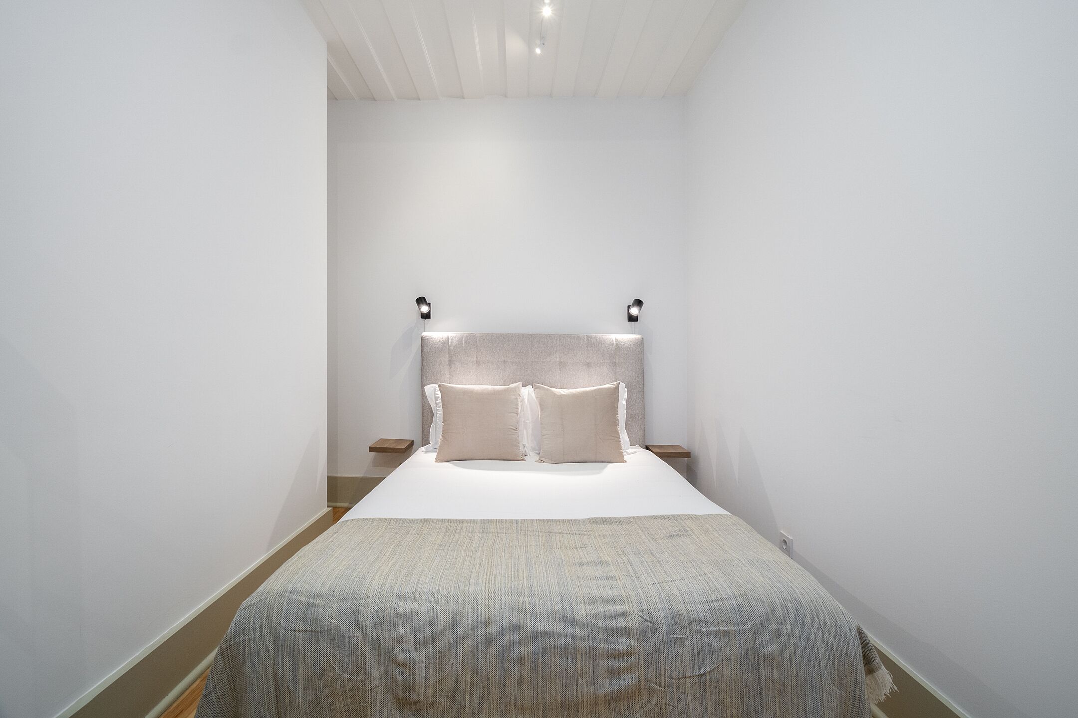 Un dormitorio minimalista con una cama, dos mesitas de noche y luces de lectura fijadas a la pared.