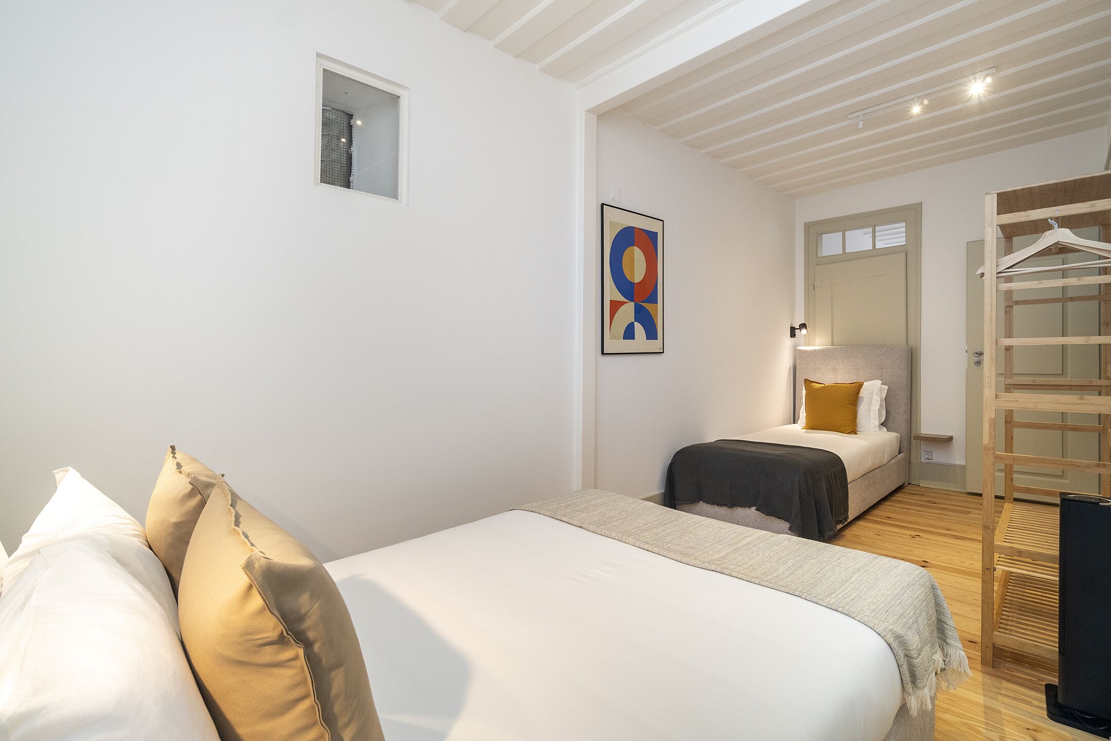 A bedroom with a bed, a small window, a wall-mounted artwork, and a wooden wardrobe.