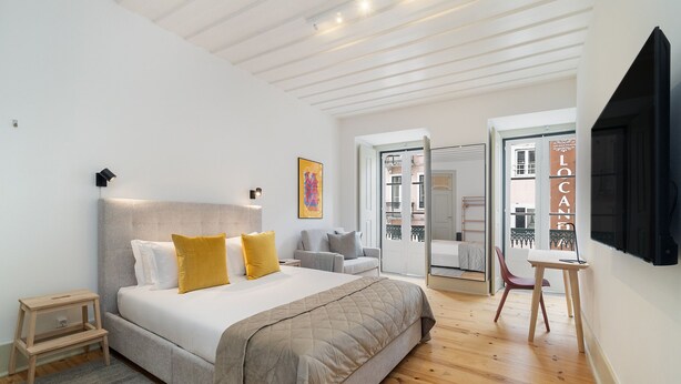 Un dormitorio con una cama grande, una mesita de noche, una silla y un escritorio. Hay una puerta que da a un balcón.
