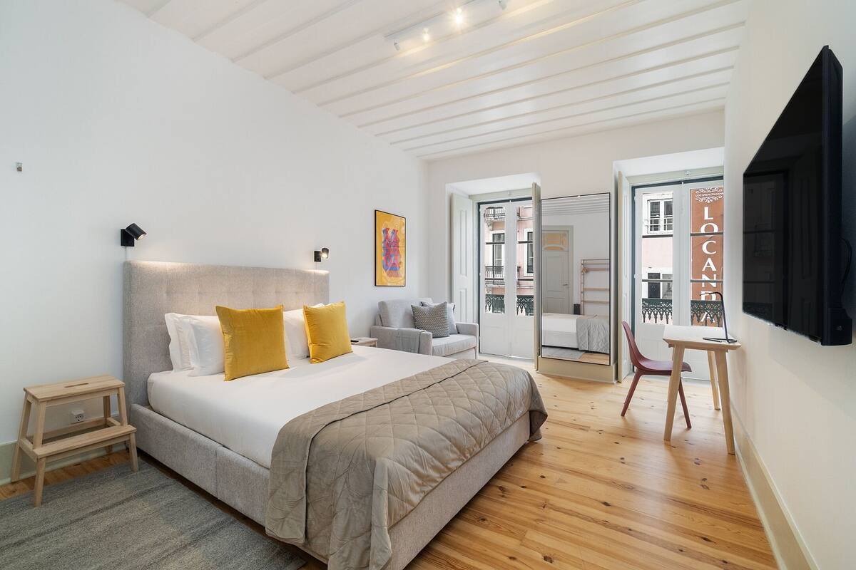 Une chambre avec un grand lit, une table de chevet, une chaise et un bureau. Une porte donne sur un balcon.