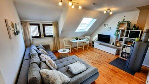 Un appartement moderne de style « loft » avec un grand canapé d’angle gris, un coin repas comprenant une petite table et des chaises, un téléviseur et un poêle à bois encastré.