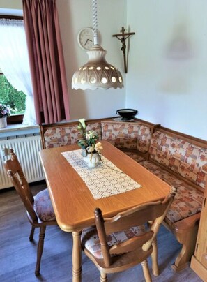 A dining area with a wooden table, two chairs, a bench, a lace table runner, a hanging light fixture, a wall clock, and a crucifix.