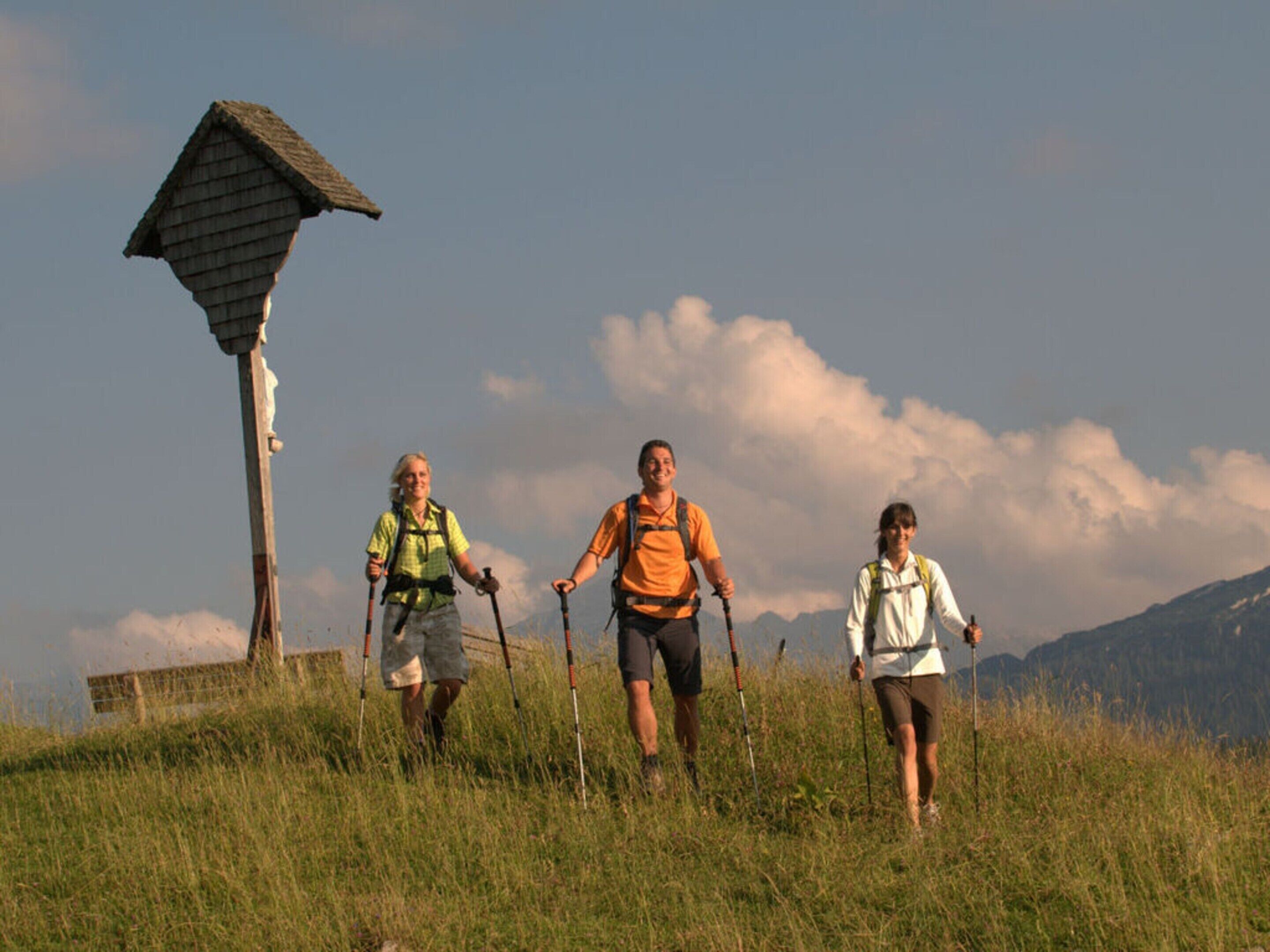 Trois personnes font de la randonnée sur une colline herbeuse, avec une structure en bois en arrière-plan.