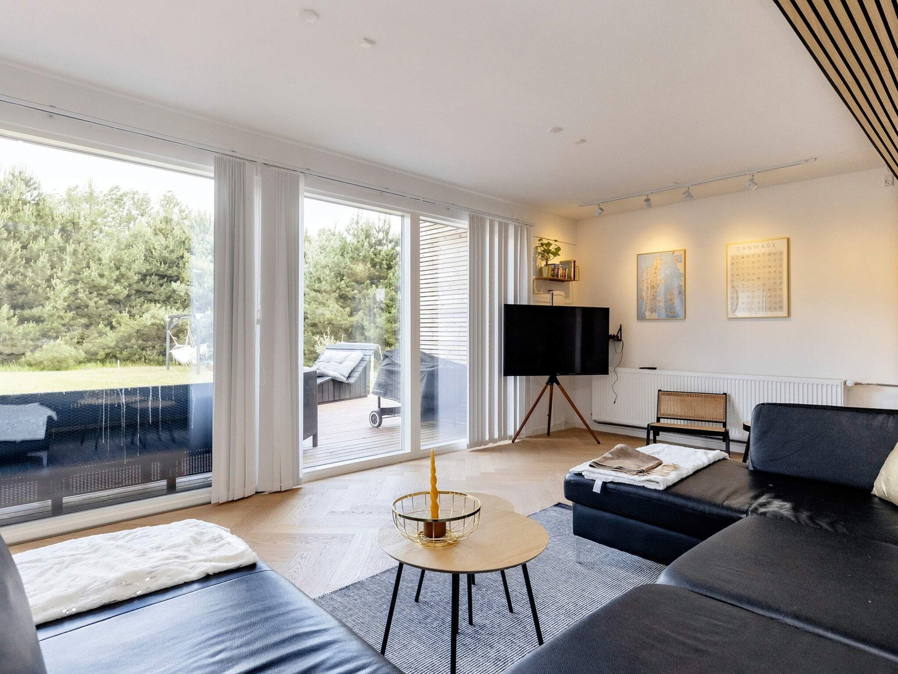 A modern living room with a large TV on a tripod, a sofa, a coffee table, and a view of a deck and trees outside.