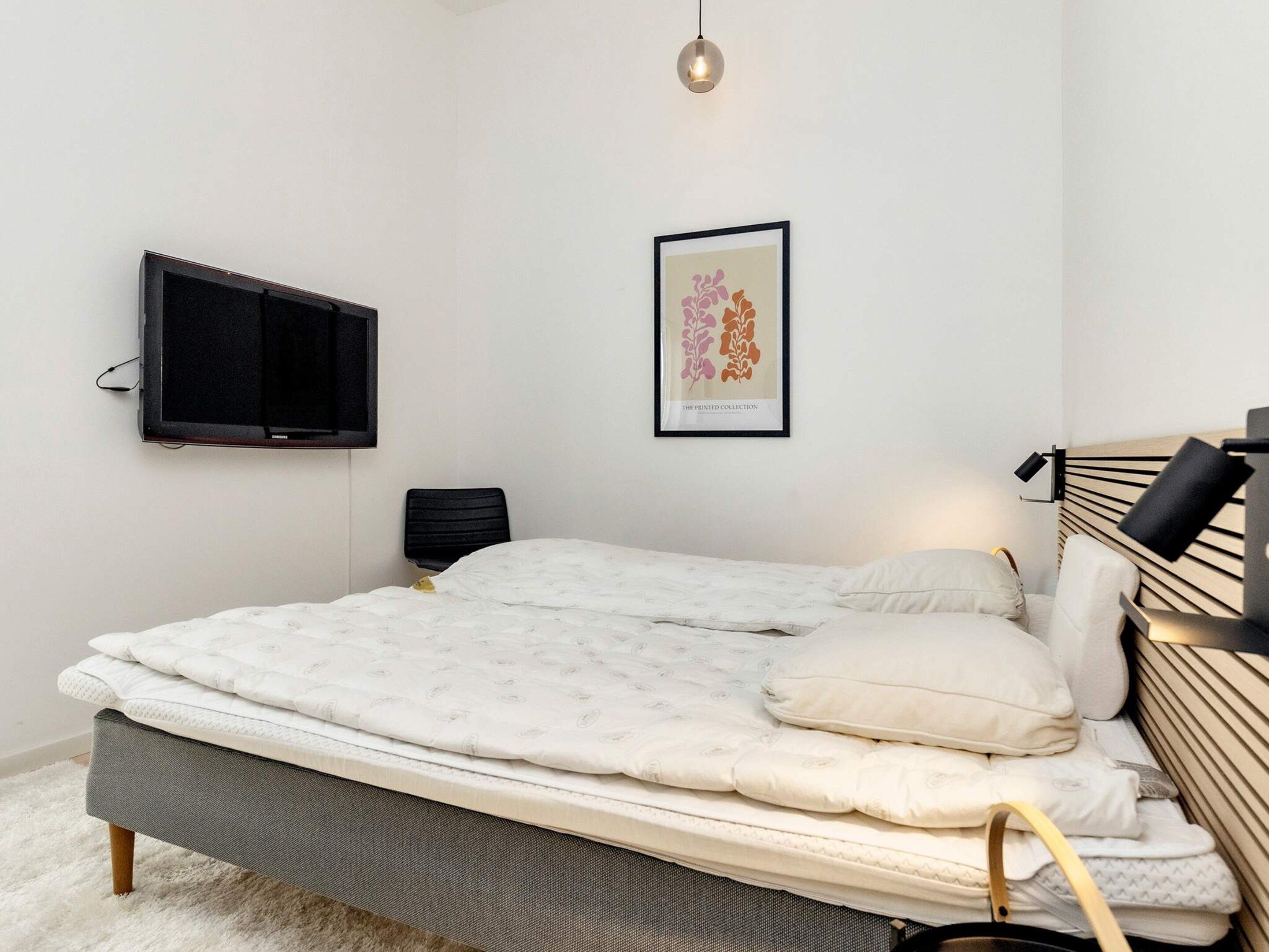 Quarto com cama, TV fixada na parede, obra de arte emoldurada e luminária de cabeceira.