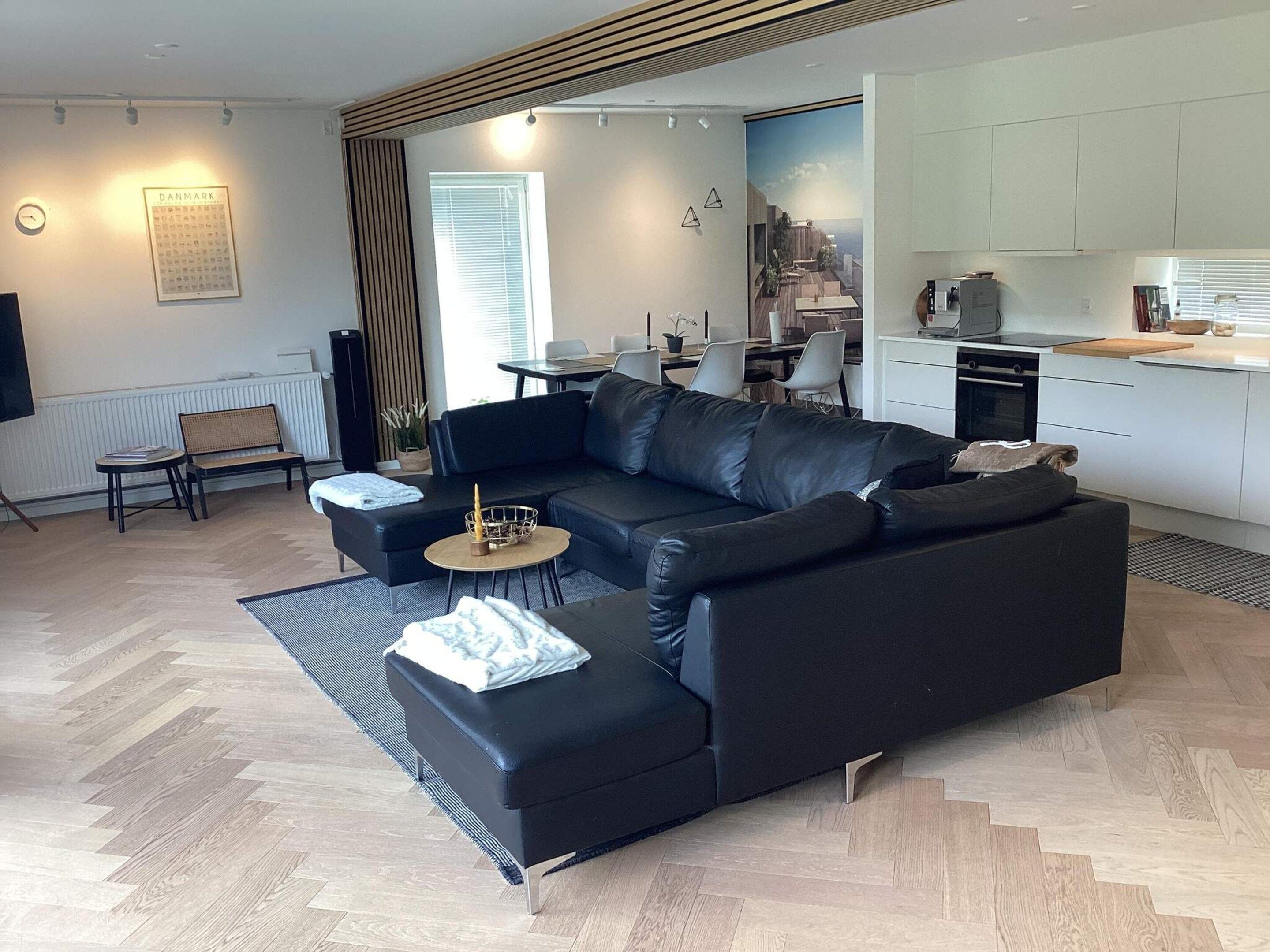 A modern living room with a black leather sofa, a round coffee table, and a dining area with a white table and chairs.
