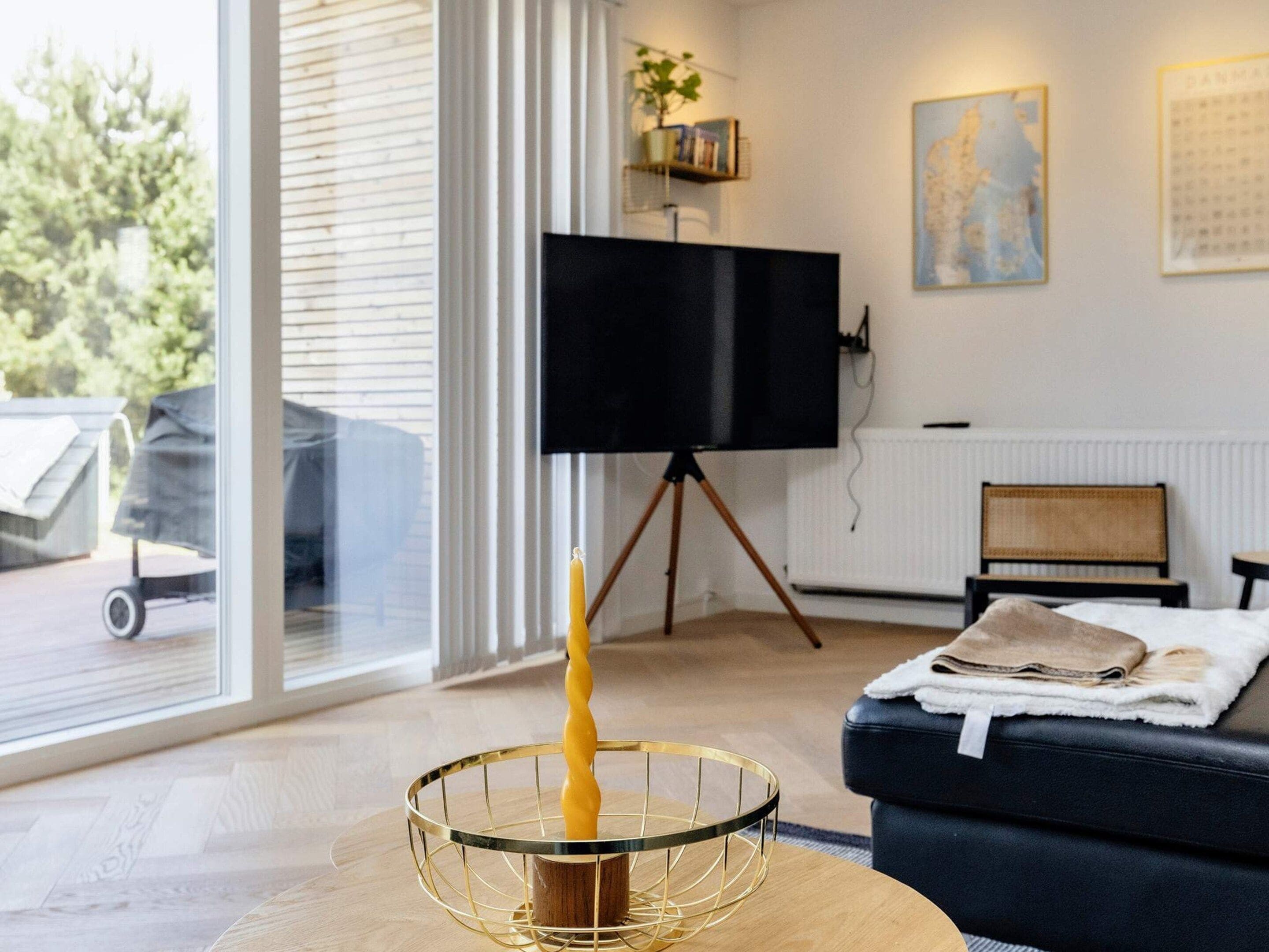A modern living room with a television on a tripod stand, a round glass table with a candle, a black sofa, and a window with blinds.